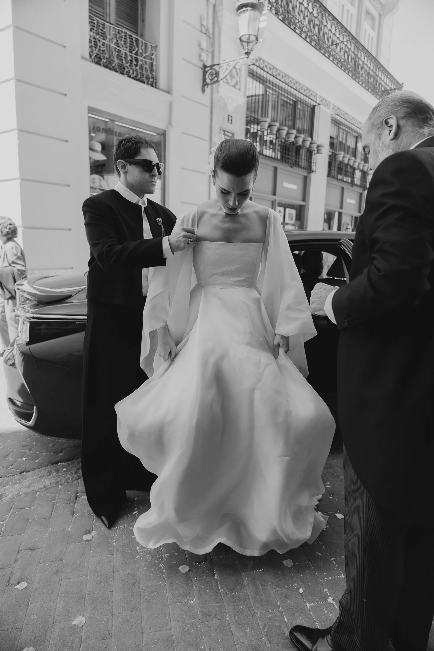 El diseñador ajusta la capa de la novia al bajar del coche, con la falda en pleno vuelo, a punto de entrar en la iglesia